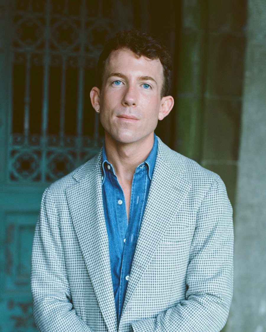 Portrait of Doyle D. Calhoun in a houndstooth jacket and denim shirt, standing before a weathered green iron gate